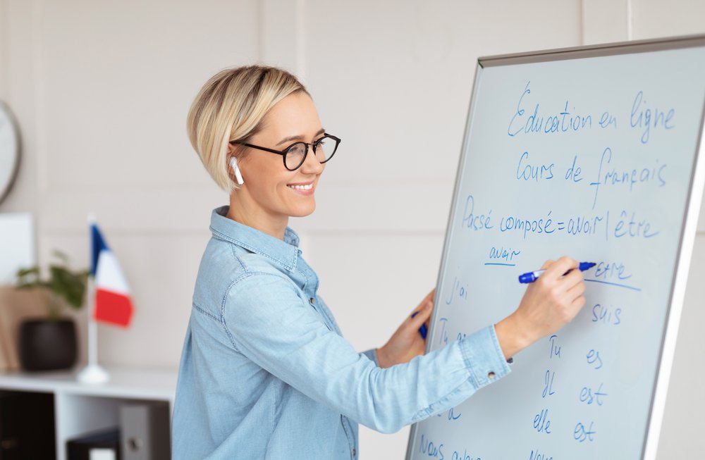 Friendly Female Teacher Writing Grammar Rules On Blackboard During Online French Lesson. Remote Education And Tutoring