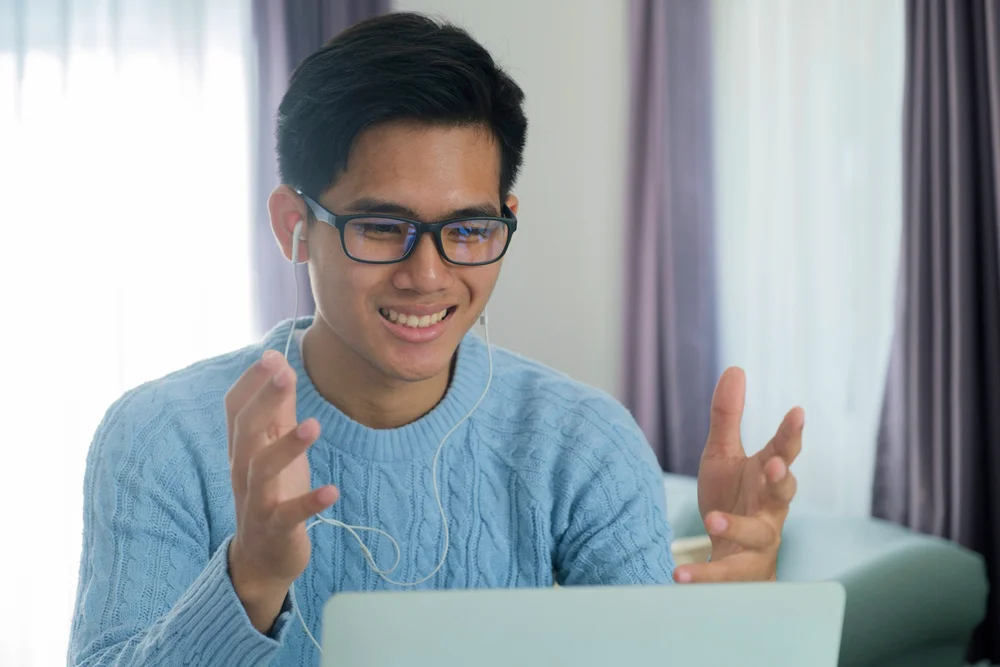 Happy young man video conferencing on laptop, wearing glasses and blue sweater.