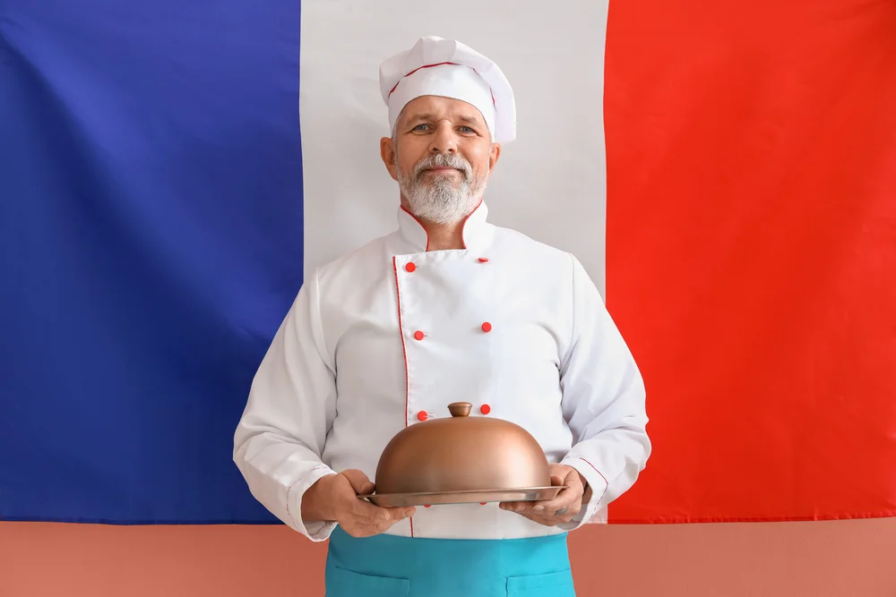 Senior french chef presents a dish under a cloche against a french flag.