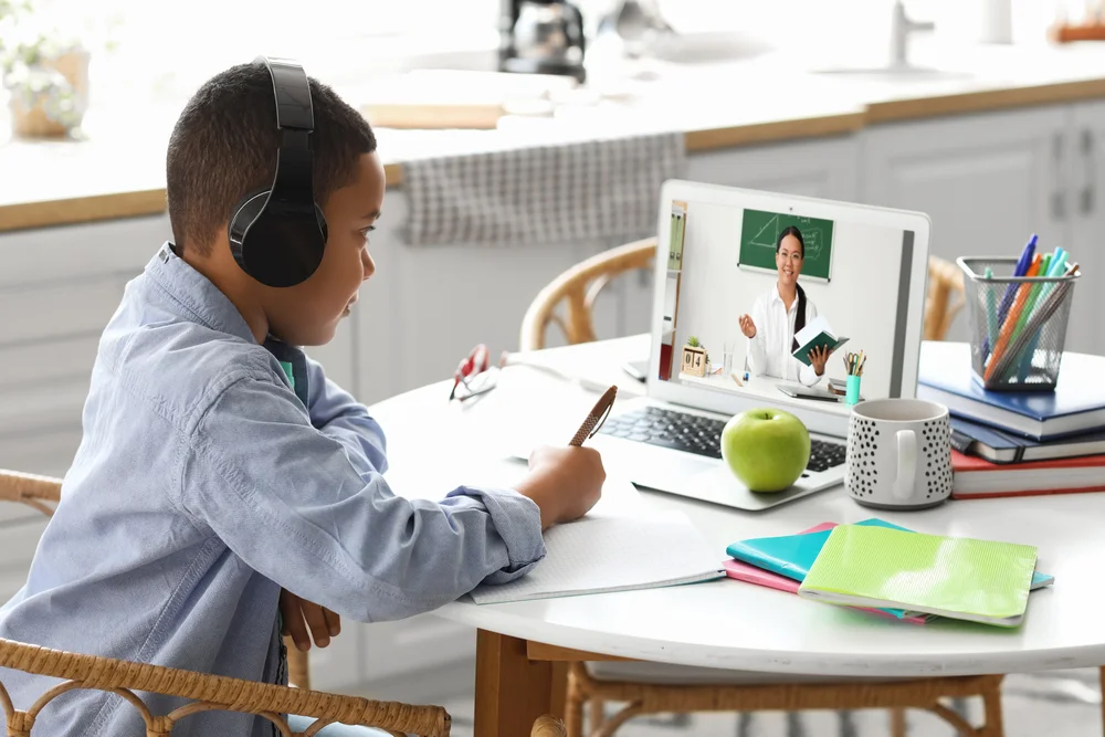Boy in headphones participates in online class via laptop, taking notes.